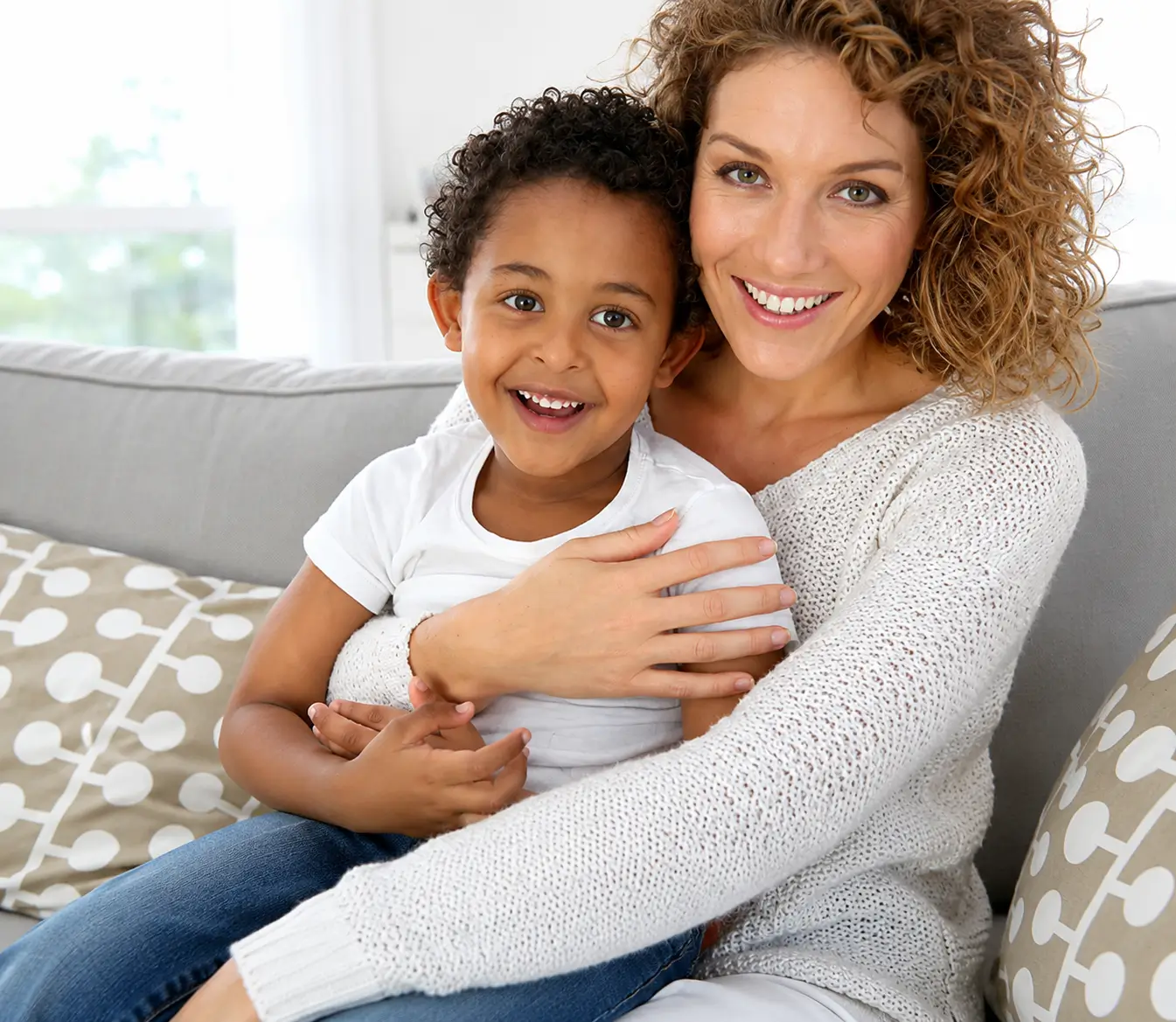 Mother embracing a smiling young child while sitting together on a couch in a bright living room.