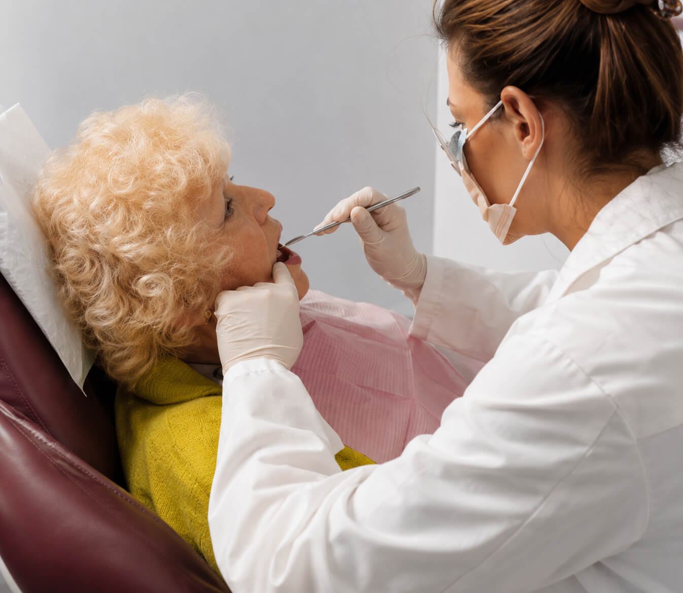 senior-dental-exam-treatment Elderly woman receiving a dental examination from a dentist wearing gloves and a face mask.